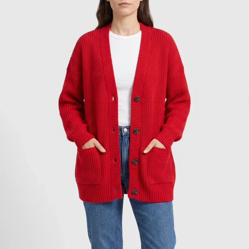 Woman wearing a long red ribbed knit cardigan with dark buttons and two front pockets, hands tucked inside.