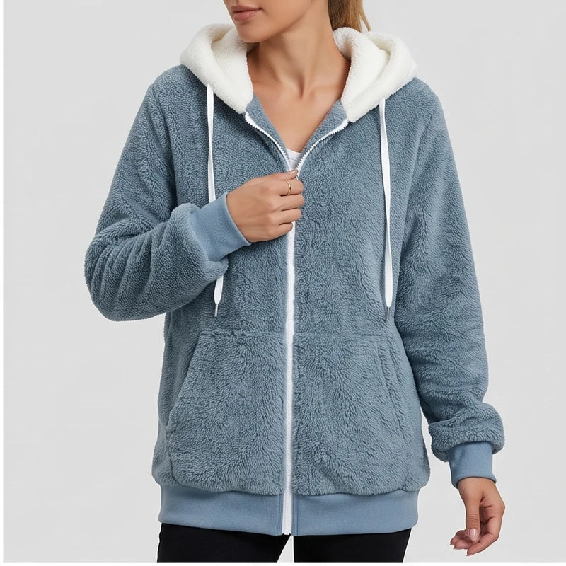 Woman zipping up a cozy blue fleece hoodie with a white sherpa-lined hood, white drawstring, and front pockets.