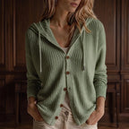 Woman wearing a light green waffle knit hooded cardigan with brown buttons, hands in pockets, against a dark wooden wall.
