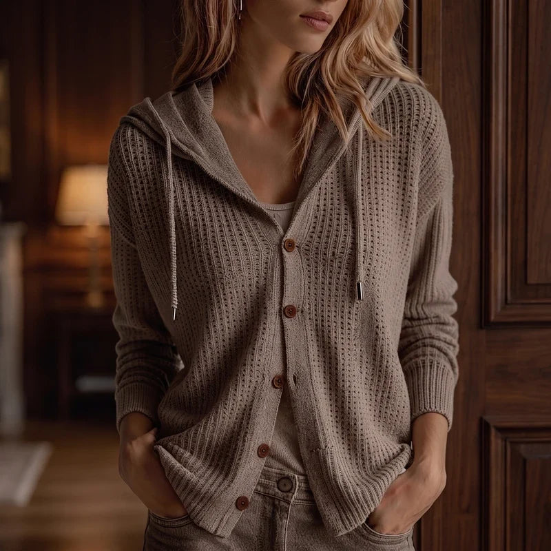 Woman wearing a light brown open-knit hooded cardigan with brown buttons, hands in pockets, in a softly lit room.
