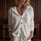 Woman wearing a relaxed-fit, cream knit hooded cardigan with brown buttons and drawstrings, hands in light denim pockets.
