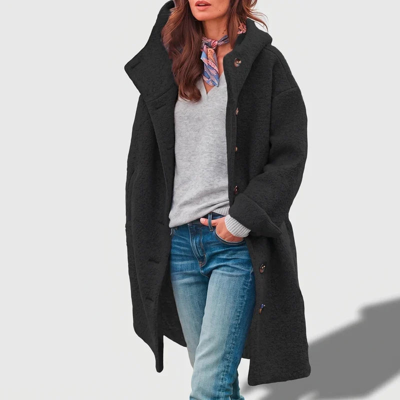 Woman wearing an open, dark-charcoal textured long hooded coat over a gray sweater and blue jeans, standing casually.