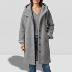 Woman wearing a long grey teddy coat with a hood, dark brown buttons, and striped cuff lining, over a grey sweater and jeans.
