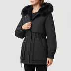Woman wearing a black parka coat with a faux fur-trimmed hood, cinched waist, and large front pockets.