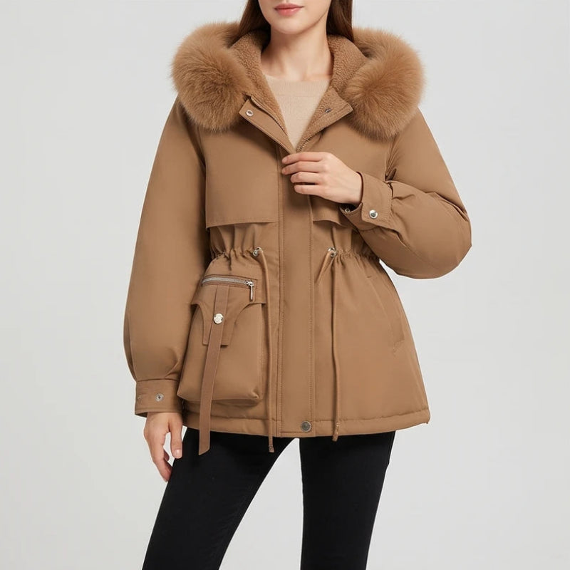 Woman wearing a tan parka coat with a faux fur-trimmed hood, drawstring waist, and flap pockets on a light background.