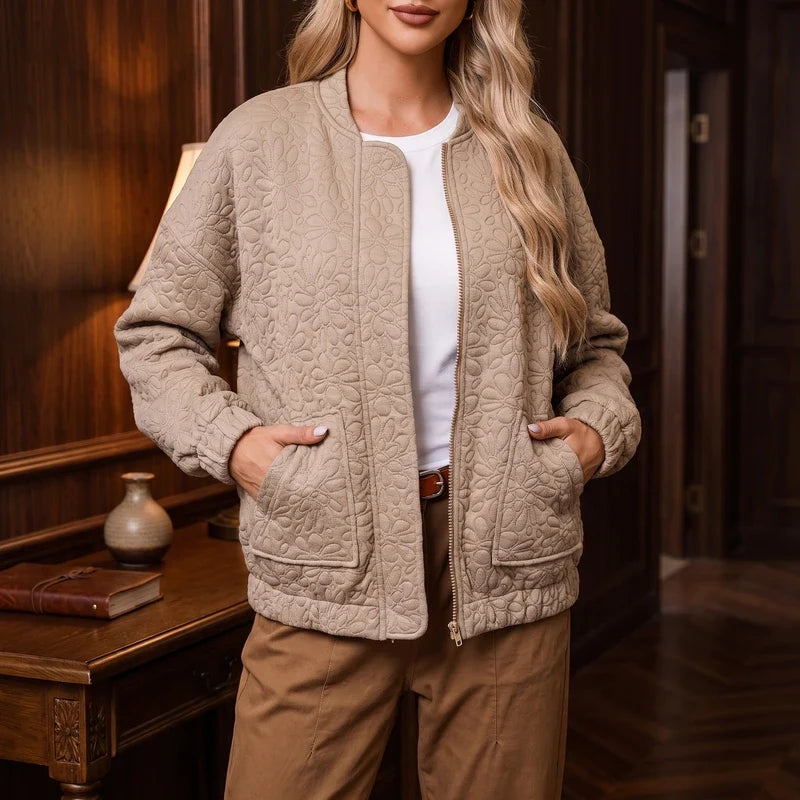 Woman wearing a beige quilted bomber jacket with a floral pattern, open over a white shirt and brown pants, hands in pockets.