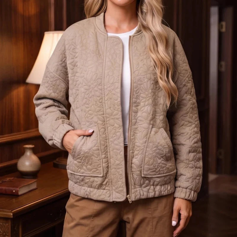 Woman wearing a beige quilted bomber jacket with floral pattern, zippered front, and elastic cuffs, standing indoors.