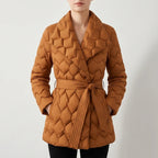 Woman wearing a rust-colored quilted puffer coat with a wide collar, tied at the waist, hands in pockets.