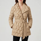 Woman wearing a beige quilted puffer coat with a wide lapel collar, tied belt, and hands in pockets on a plain white background.