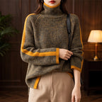 Woman wearing a dark green marl roll neck knit jumper with wide orange stripes on the sleeves and hem over light brown pants.