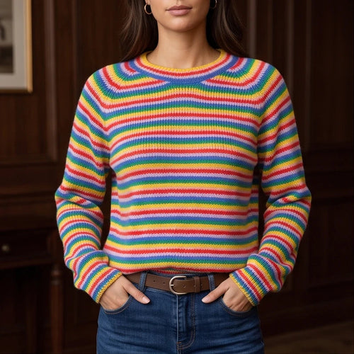 Woman wearing a rainbow-striped knit jumper with long sleeves, blue jeans, and a brown belt, hands in pockets.