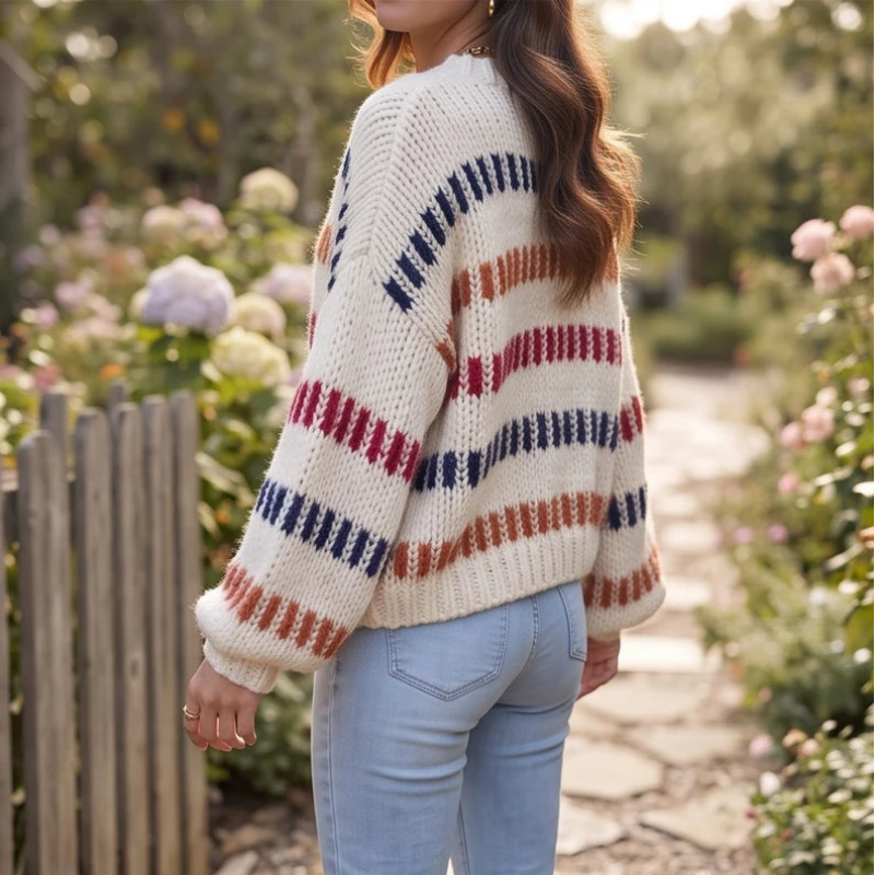 Woman wearing a cream chunky knit sweater with colorful horizontal stripes, light wash jeans, standing in a garden.