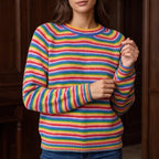 Woman wearing a rainbow-striped knit jumper with a crew neck and ribbed cuffs, against a blurred wooden background.