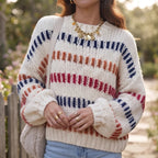 Woman wearing a cream chunky knit jumper with horizontal red, orange, and navy striped patterns, outdoors.
