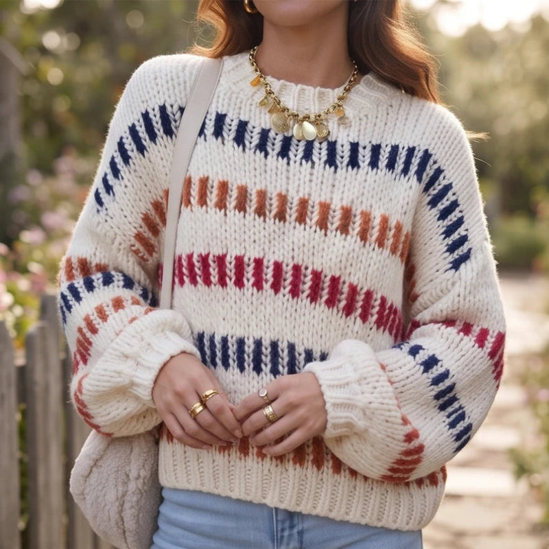 Woman wearing a cream chunky knit jumper with horizontal red, orange, and navy striped patterns, outdoors.