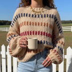 Woman wearing a cream chunky knit sweater with blue, orange, and red striped patterns, holding a light beige mug outdoors.