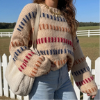 Woman wearing a chunky beige knit jumper with colorful red, blue, and orange stripes, standing outdoors by a white fence.
