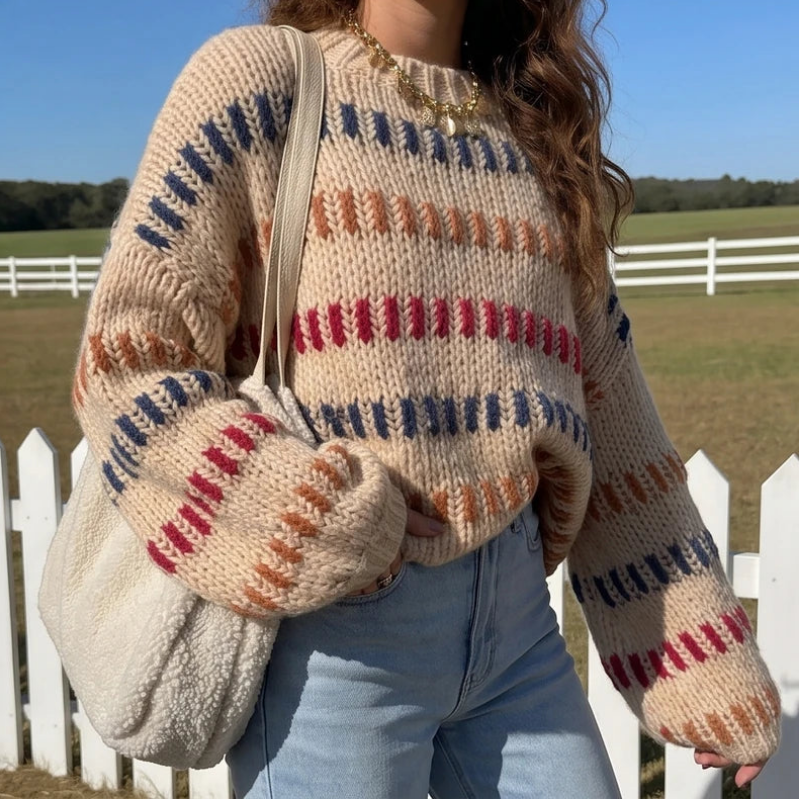 Woman wearing a chunky beige knit jumper with colorful red, blue, and orange stripes, standing outdoors by a white fence.