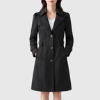 Woman wearing a black knee-length trench coat with puffed sleeves, three brown buttons, and hands in side pockets.