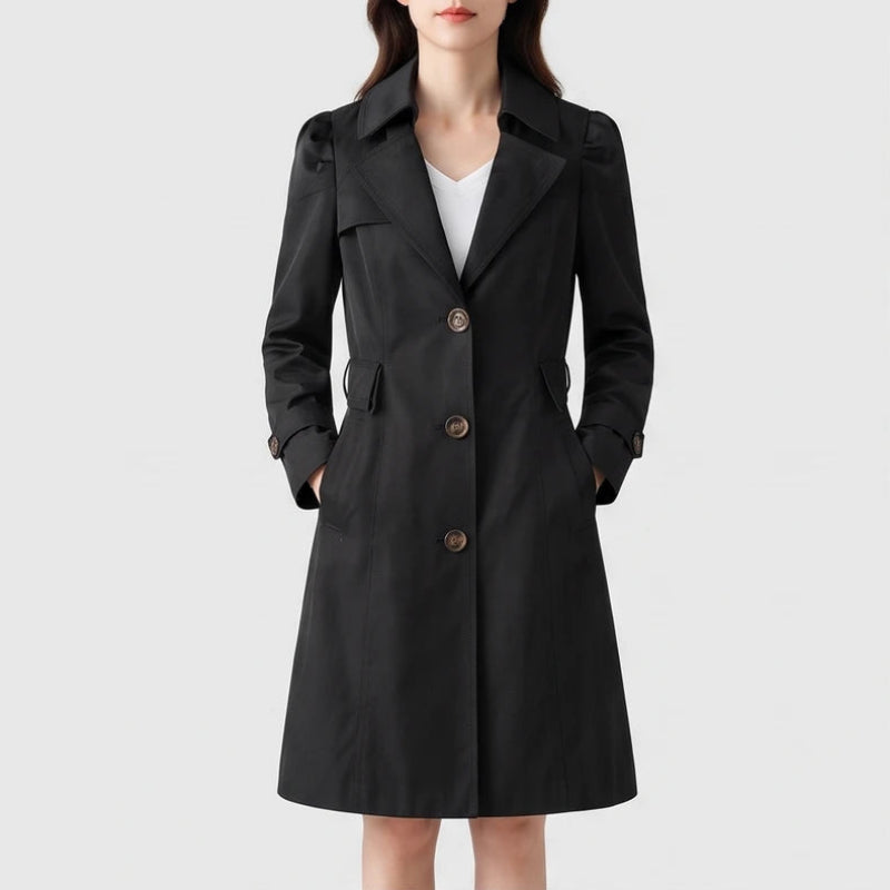 Woman wearing a black knee-length trench coat with puffed sleeves, three brown buttons, and hands in side pockets.