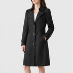 Woman wearing a black trench coat with large brown buttons, a structured collar, and belted cuffs, with hand in pocket.
