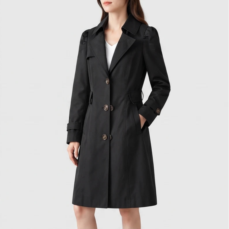 Woman wearing a black trench coat with large brown buttons, a structured collar, and belted cuffs, with hand in pocket.