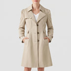 Woman wearing a beige trench coat with puff shoulders, brown buttons, and hands in the side pockets, against a white background.